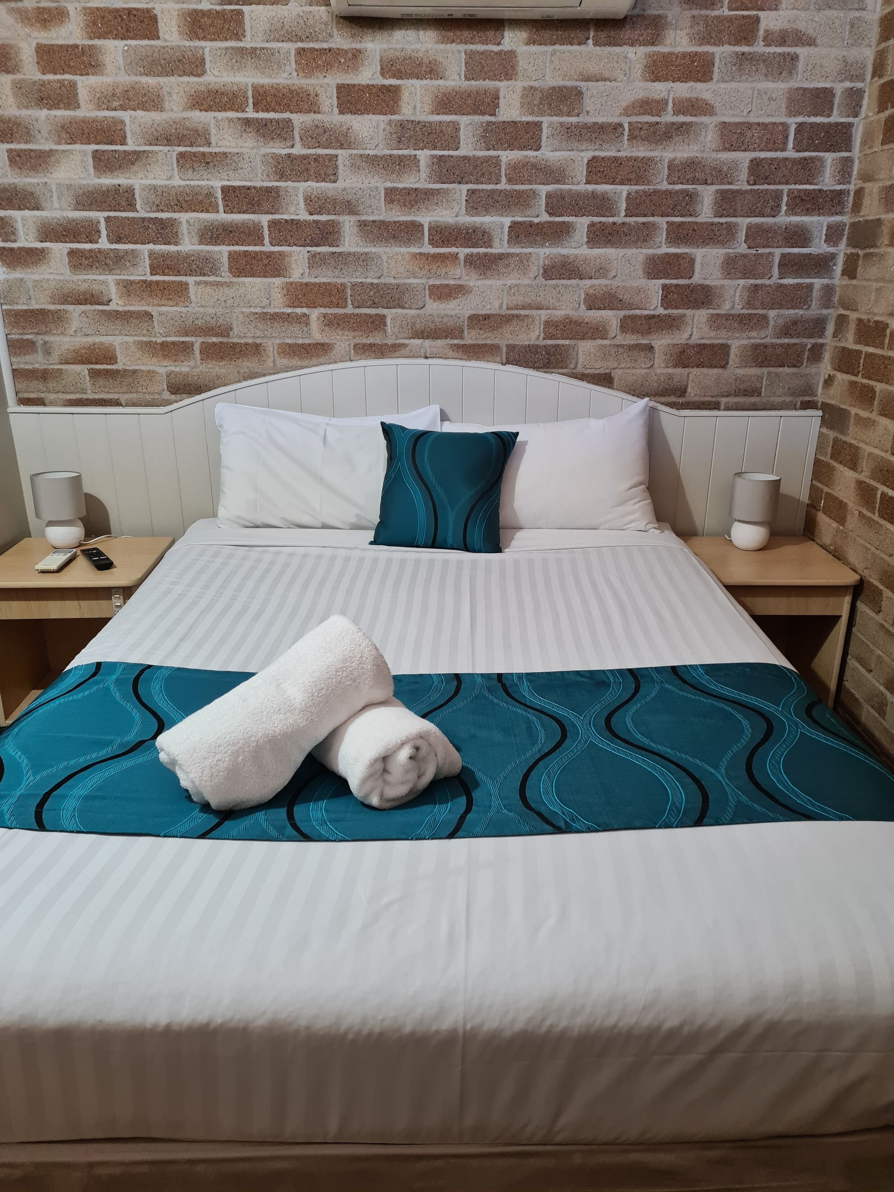 Un lit bien fait, avec une literie blanche et turquoise, un oreiller blanc et une serviette blanche enroulée, posés sur une couvre-lit bleue à motifs. On aperçoit deux tables de chevet avec des lampes, et un mur en briques en arrière-plan.