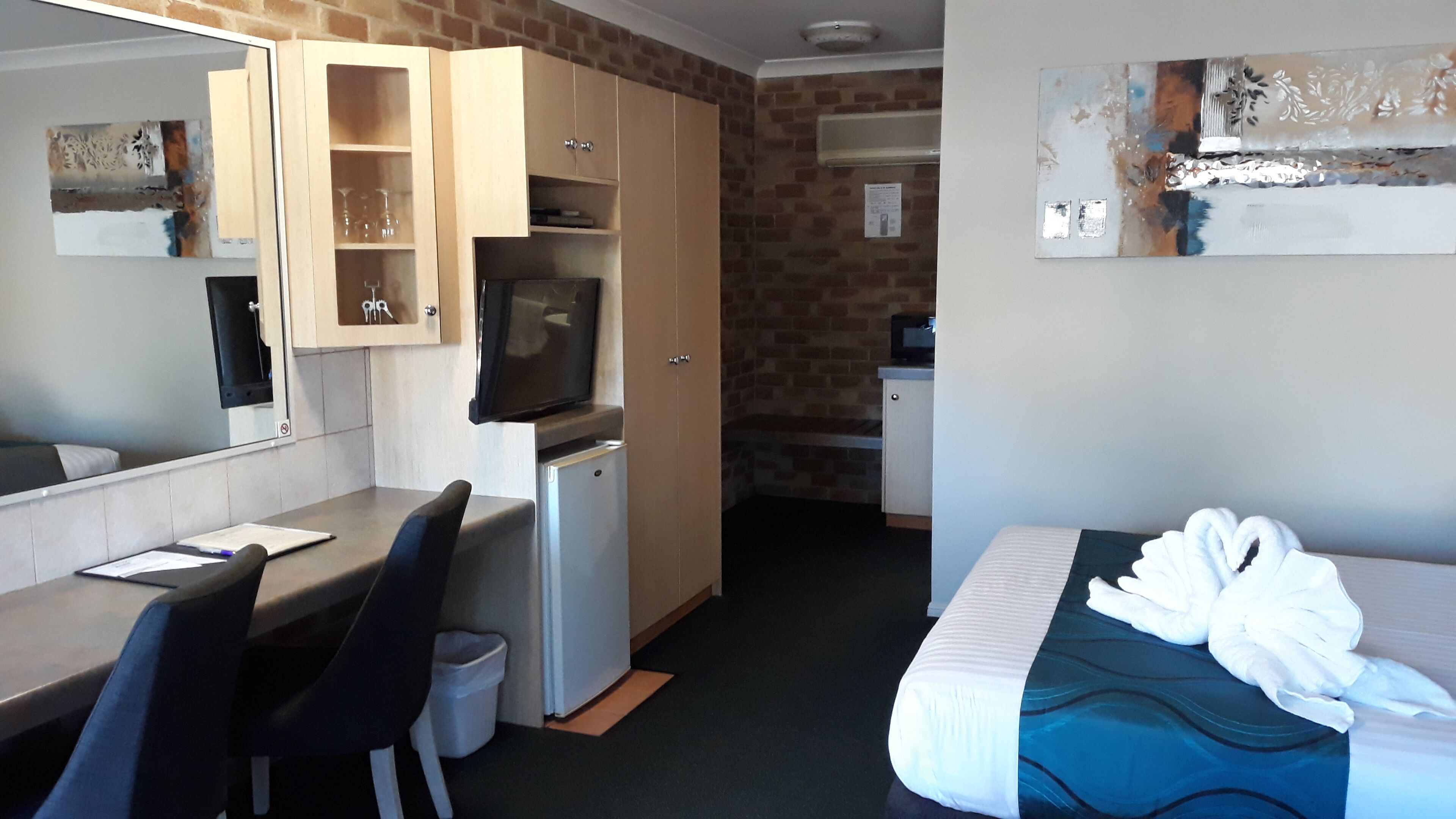 Une chambre d’hôtel compacte comprenant un coin cuisine, un lit agrémenté d’une serviette en forme de cœur, un bureau avec une chaise, une télévision et un miroir.