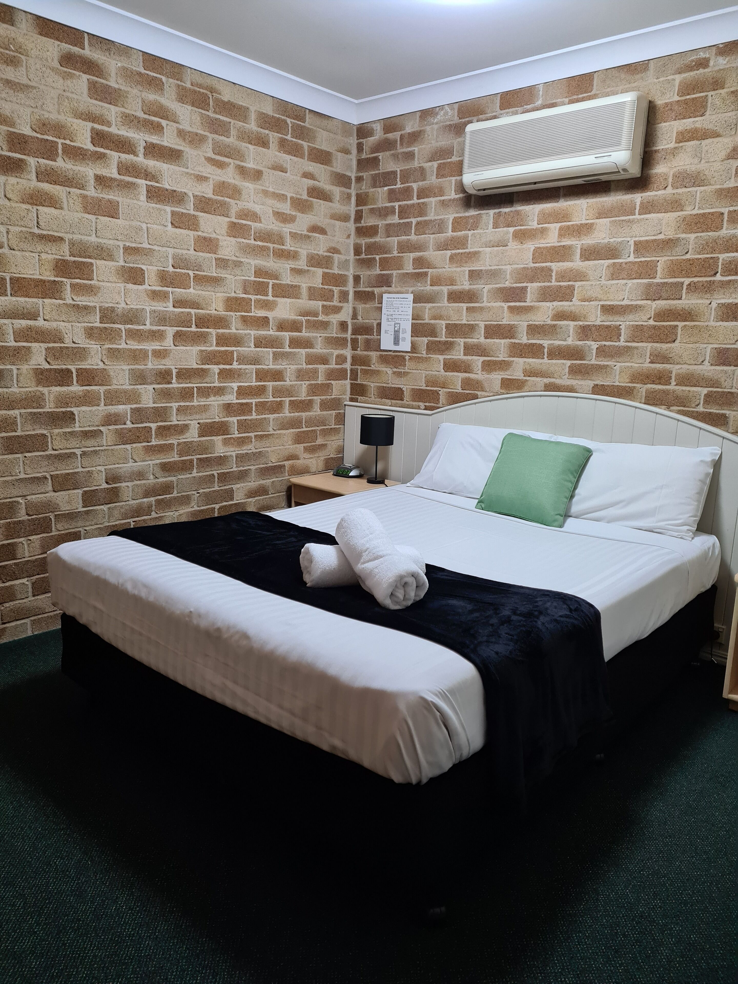 Una habitación de hotel con pared de ladrillo, cama con ropa de cama blanca y verde, manta negra, mesita de noche con lámpara y aire acondicionado de pared.