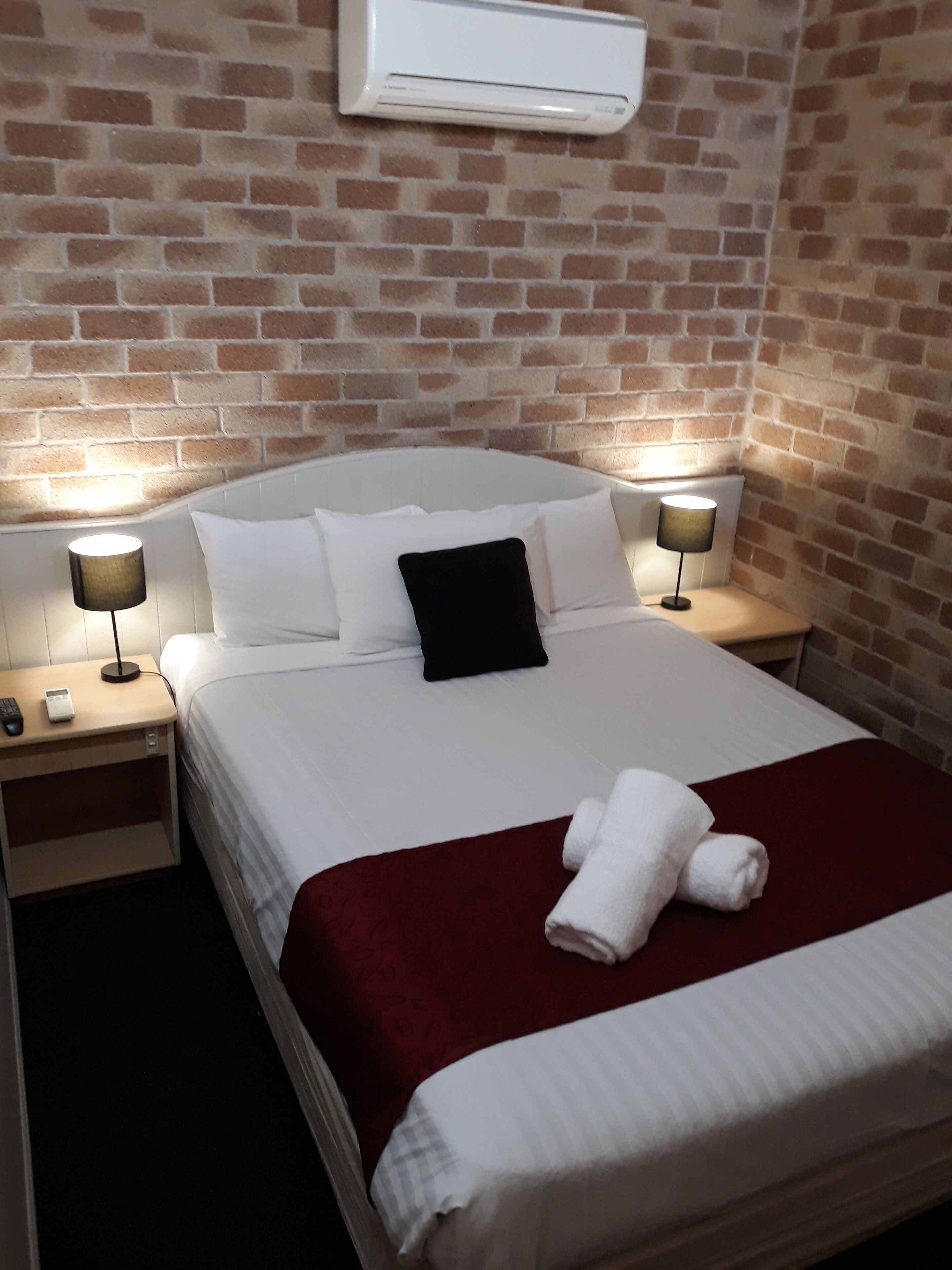 A hotel room with a brick wall, a white bed with a black pillow, two bedside lamps, and a red throw blanket.