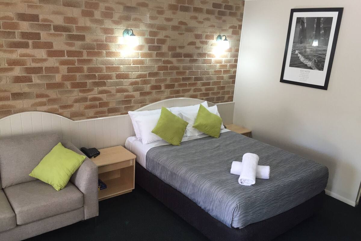 A hotel room with a brick wall, a bed with a grey blanket and pillows, a small bedside table with a phone, and a framed picture on the wall.