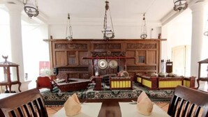A dining area with a wooden table set for two, surrounded by traditional patterned sofas and a decorative wall with a clock. - Roemahkoe Heritage Hotel (Surakarta)