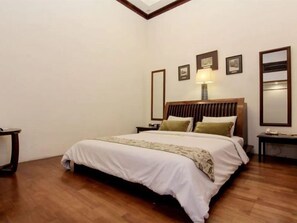 A hotel room with a large bed, wooden flooring, a nightstand with a lamp, and framed pictures on the wall. - Roemahkoe Heritage Hotel (Surakarta)