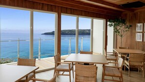 Ein geräumiges Zimmer mit großen Fenstern, das Meer- und eine entfernte Inselblick bietet. Holztafeln und Korbstühle sind zum Essen angeordnet.