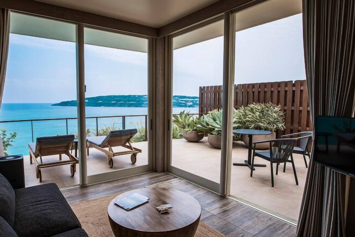 Een balkon met uitzicht op zee, een bank, een salontafel en een eethoek met stoelen en planten.