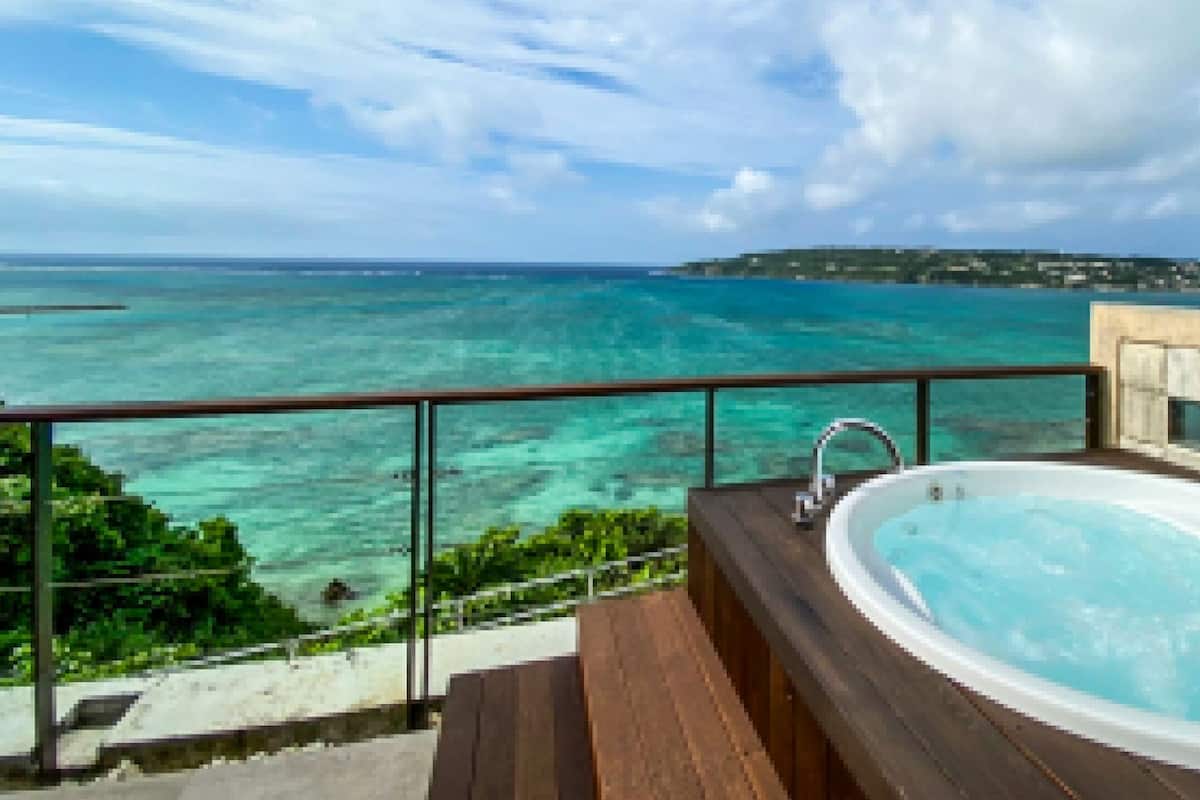 Een hottub op een houten terras met glazen balustrade, uitkijkend op een turquoise zee en een eiland in de verte.