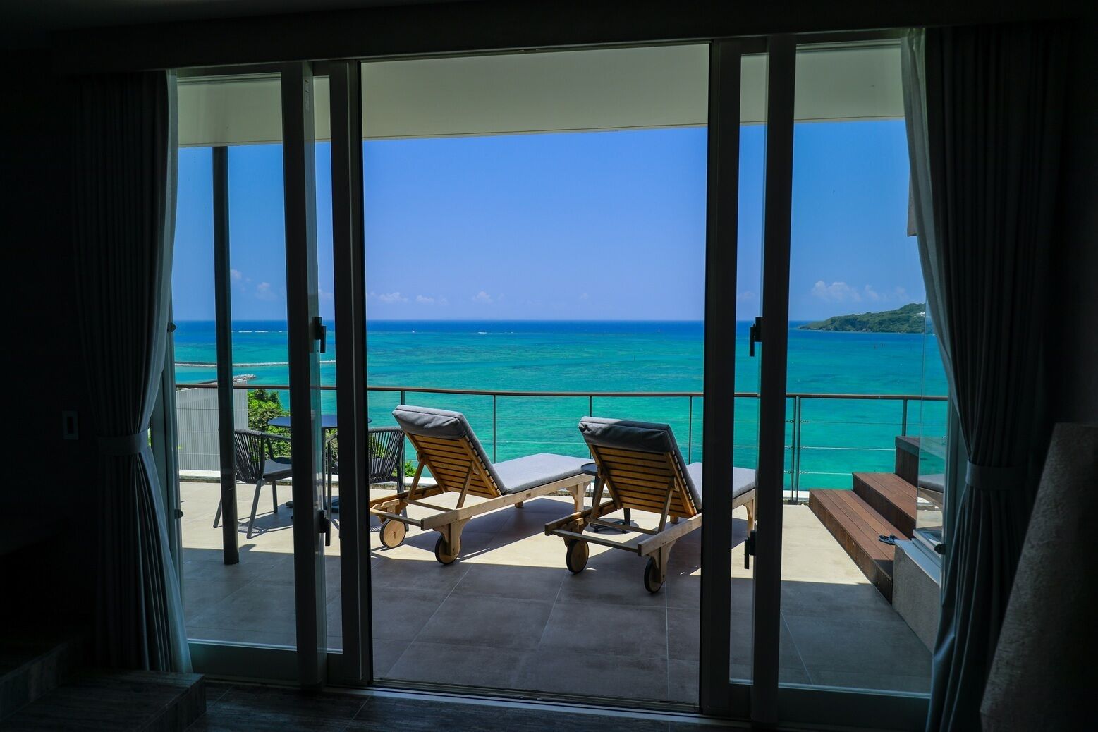 Een balkon met twee loungesteunen en een tafel, met uitzicht op een turquoise oceaan en een heldere lucht.