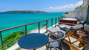Ein Balkon mit Blick auf türkisfarbenes Wasser und eine entfernte Insel. Zwei runde Tische und eine hölzerne Bank mit Kissen sind vorhanden.