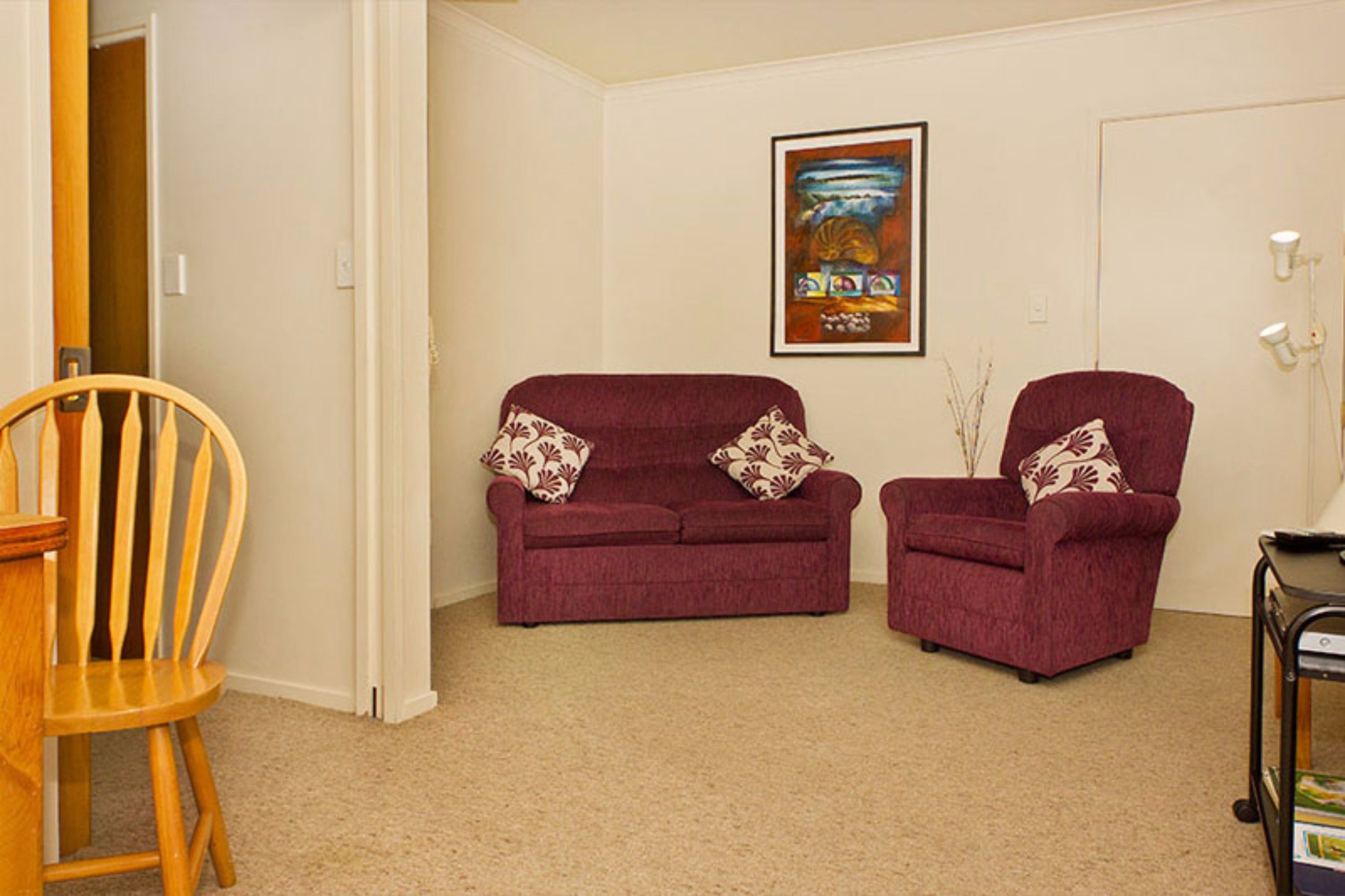 Ein Wohnzimmer mit einem Holzstuhl, einem roten Sofa und einem roten Sessel. An der Wand hängt ein gerahmtes Bild und im Hintergrund ist eine Tür zu sehen.