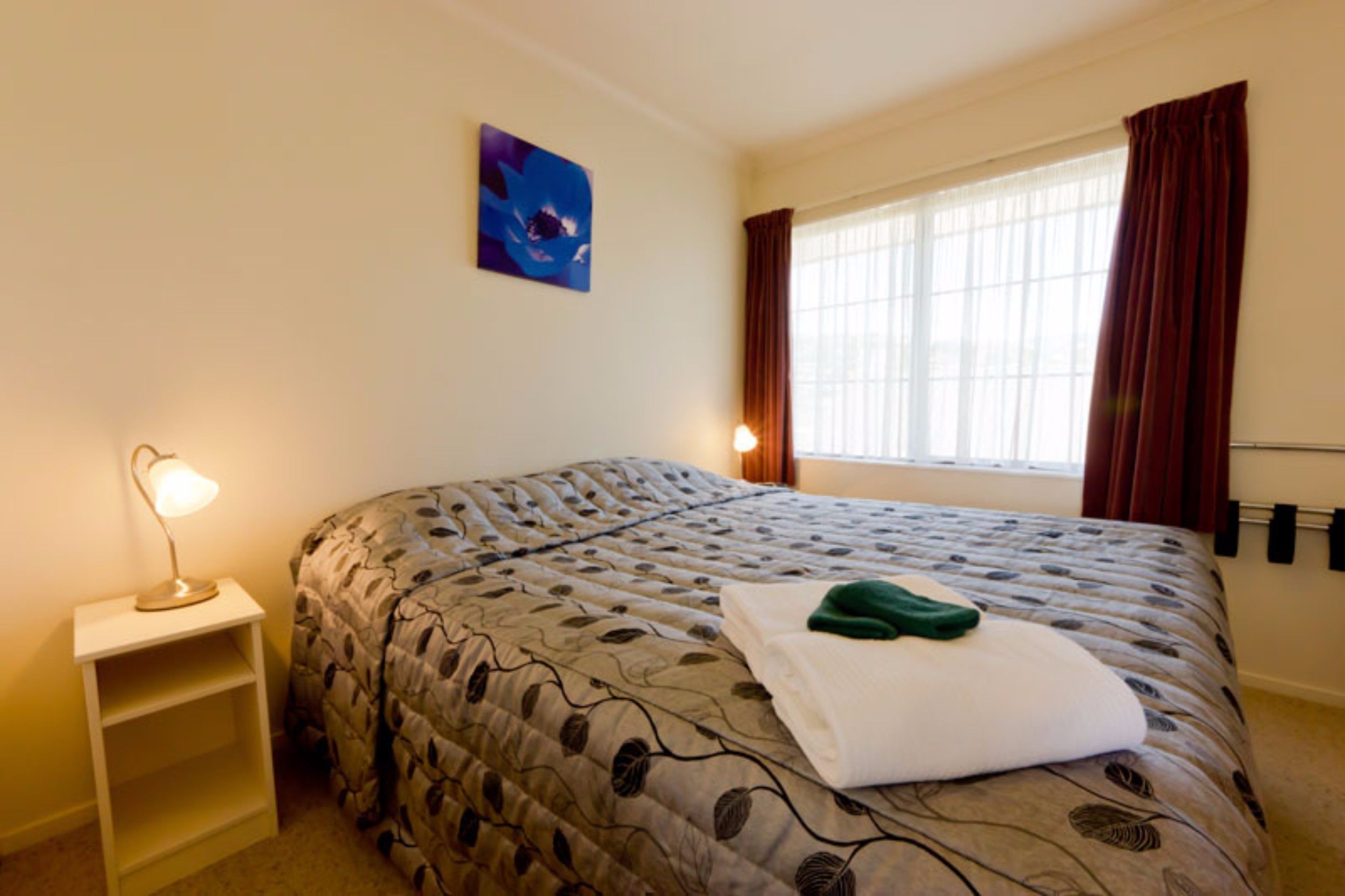 A neatly made bed with a leaf-patterned duvet, a bedside lamp, a painting on the wall, and a window with curtains.