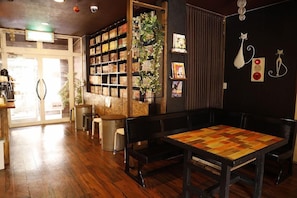 A cozy cafe with wooden flooring, a central table with benches, a bookshelf filled with books, and a decorative wall with cat figurines.