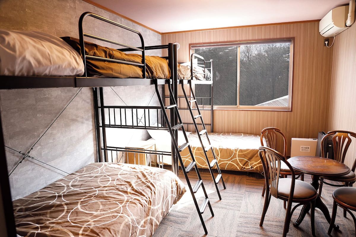 A dormitory room with bunk beds, a desk, chairs, and a window.