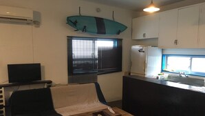 A compact living space with a sofa, a coffee table with remote controls, a surfboard decoration, a kitchen area with a refrigerator, and a window with blinds. - Life Rock (Chatan)