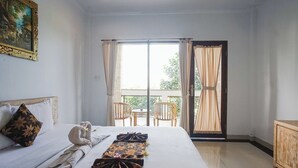 A hotel room with a bed, a painting on the wall, a balcony with chairs, and a view of greenery. - Agung Homestay Canggu (Canggu)