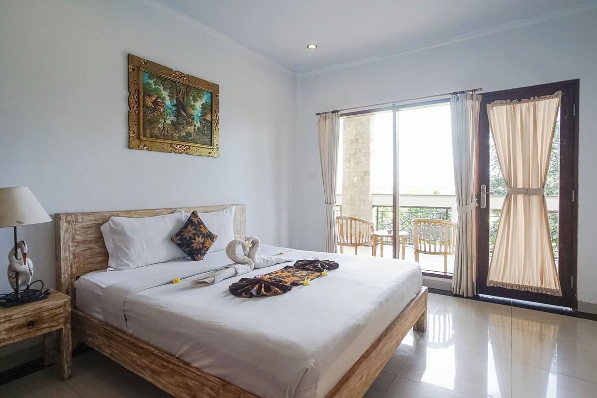 A well-lit bedroom with a large bed, a nightstand with a lamp, a framed painting on the wall, and a balcony with curtains.