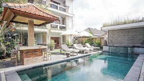 A swimming pool with lounge chairs and umbrellas, surrounded by a garden and a building with a tiled roof. - Agung Homestay Canggu (Canggu)