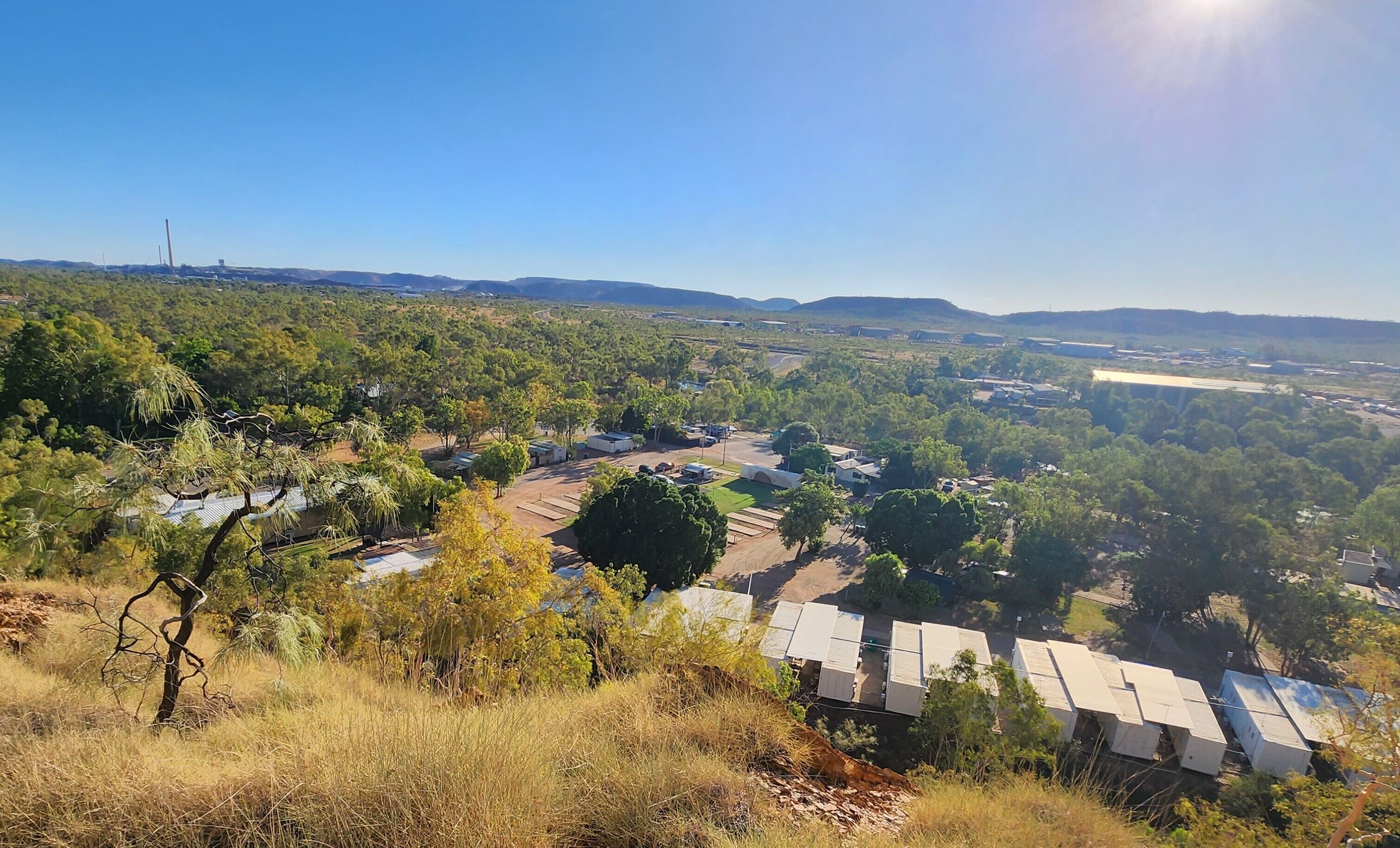 A rural landscape with scattered buildings, including a row of cabins with flat roofs, surrounded by trees and open fields.