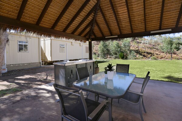 An outdoor patio with a glass table and metal chairs, a barbecue grill, and a thatched roof structure. - Aaok Moondarra Accommodation Village (Lanskey)