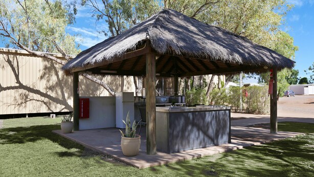 Una estructura al aire libre con techo de paja e incluye una zona de bar, rodeada de un césped cuidado y árboles.