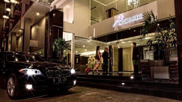 A modern hotel entrance with a glass facade, a BMW parked in front, and people walking towards the entrance. - Orchardz Ayani (Pontianak)