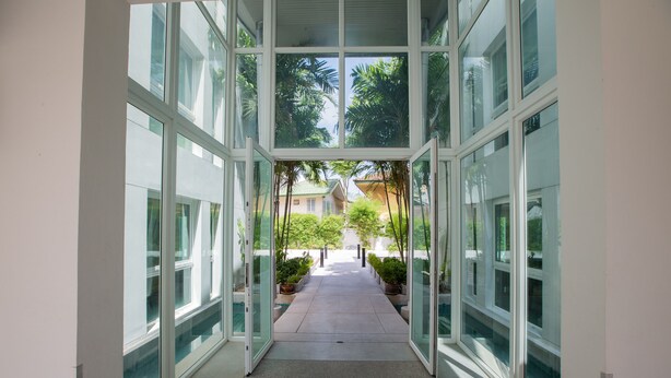 A corridor with glass walls and doors, leading to a courtyard with potted plants and a building in the background. - The Palms Kamala Beach (Kamala)