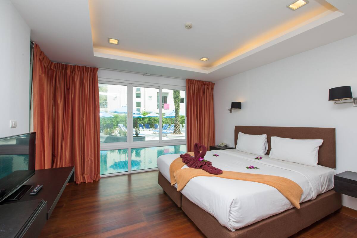 A modern hotel room with a large bed, a TV, and a view of a pool and outdoor area.