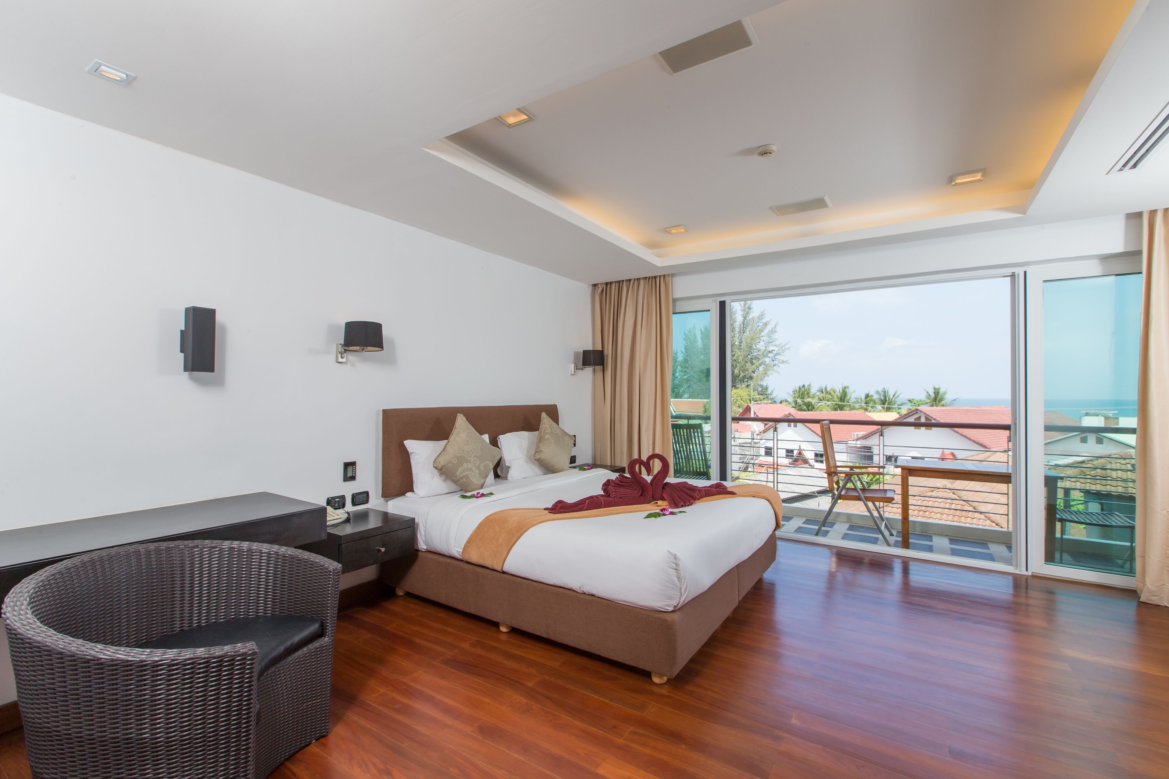 A modern hotel room with a large bed, a desk, a wicker chair, and a balcony with a view of the sea.
