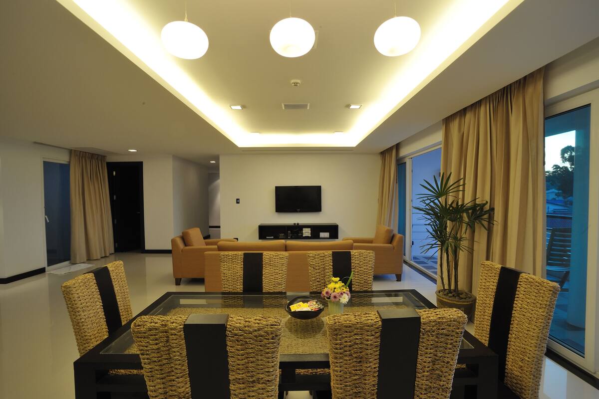 A modern living room with a sofa set, a glass-top dining table with wicker chairs, a flat-screen TV mounted on the wall, and a potted plant.