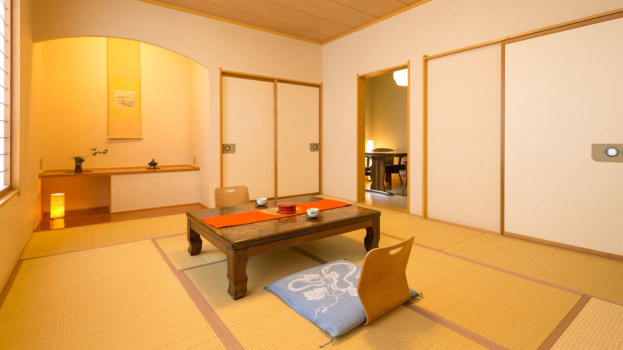 Una habitación japonesa tradicional con suelo de tatami, una mesa baja con un tapete rojo, un cojín y una puerta corredera.