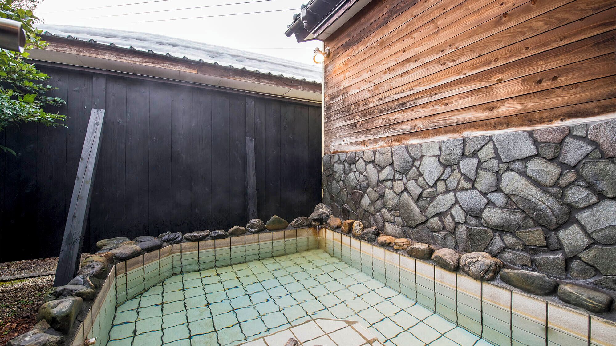 Un pequeño espacio con jacuzzi al aire libre, con suelo de baldosas, rodeado por un muro de piedra y valla de madera.