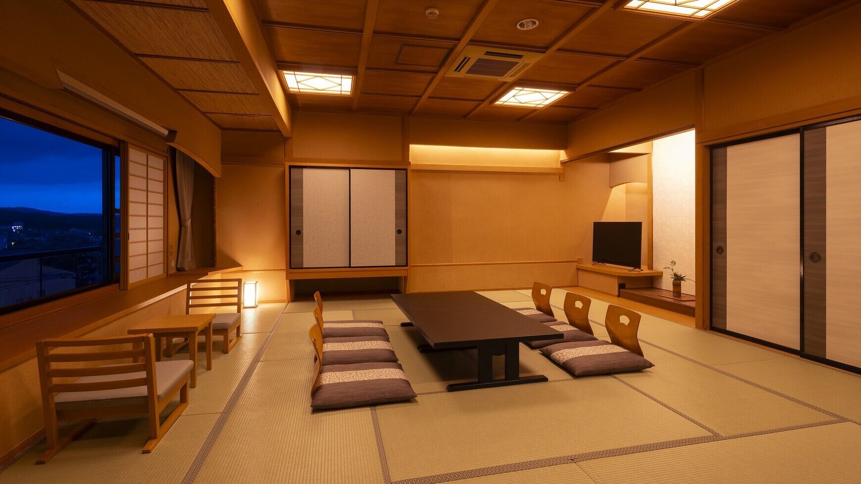 A traditional Japanese room with tatami flooring, a low table, wooden chairs, sliding doors, and a television.