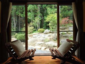 A room with two wooden lounge chairs facing a large sliding glass door leading to a garden with rocks and trees. - All Resort Service Holiday Homes and Cottages to Rent (Inawashiro)