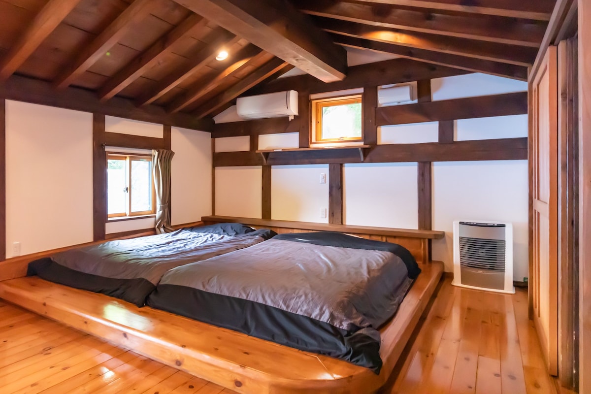 A room with a wooden floor, a large bed with a headboard, and two windows with curtains.