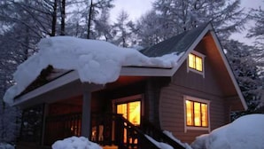 A cabin with a wooden exterior, a porch, and a visible window. Snow covers the roof and surrounding area. - All Resort Service Holiday Homes and Cottages to Rent (Inawashiro)