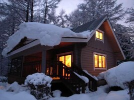 Una cabaña con fachada de madera, un porche y una ventana visible. Nieve cubre el techo y las zonas aledañas.