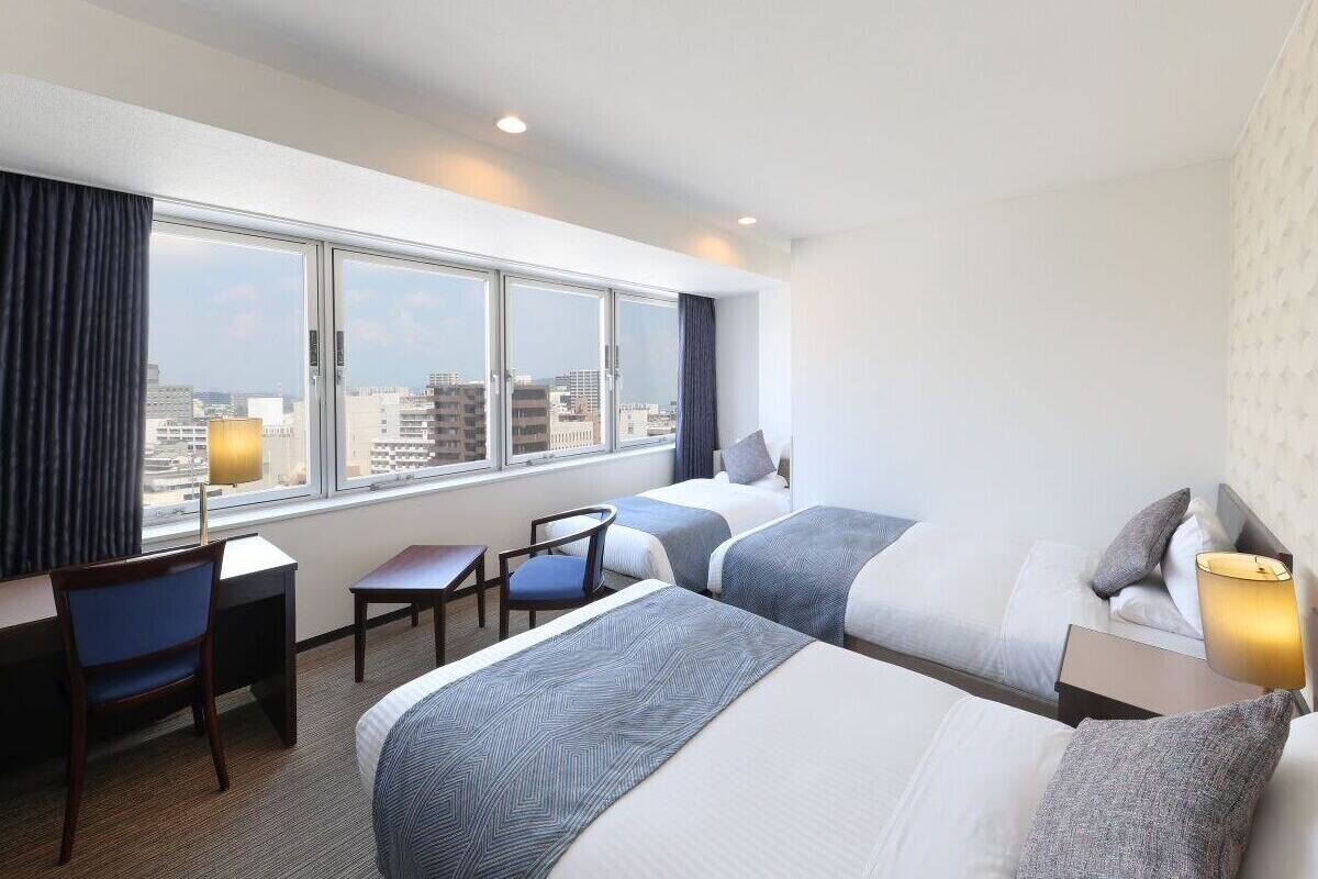 A hotel room with two beds, a desk, a chair, and a large window with a city view.