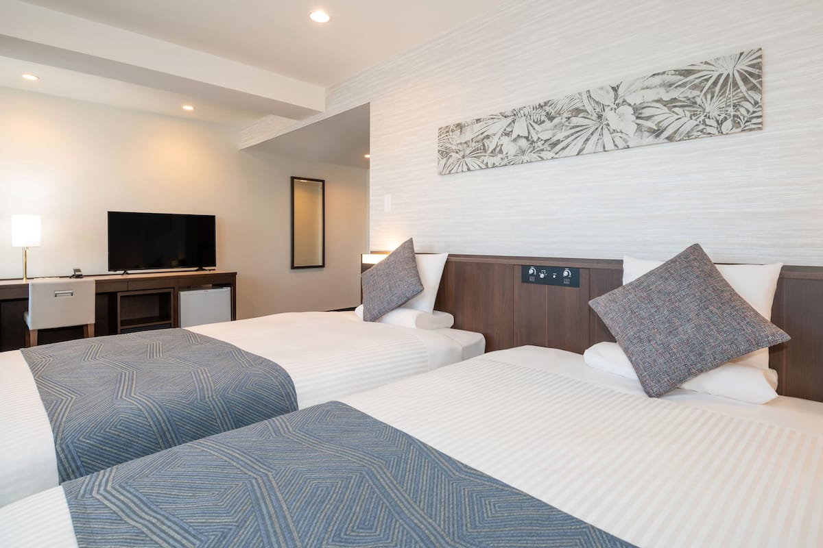 A hotel room with two beds, a desk, a TV, and a wall-mounted control panel.