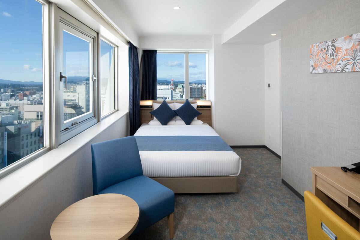 A hotel room with a bed, a seating area, a small table, and a view of the city.