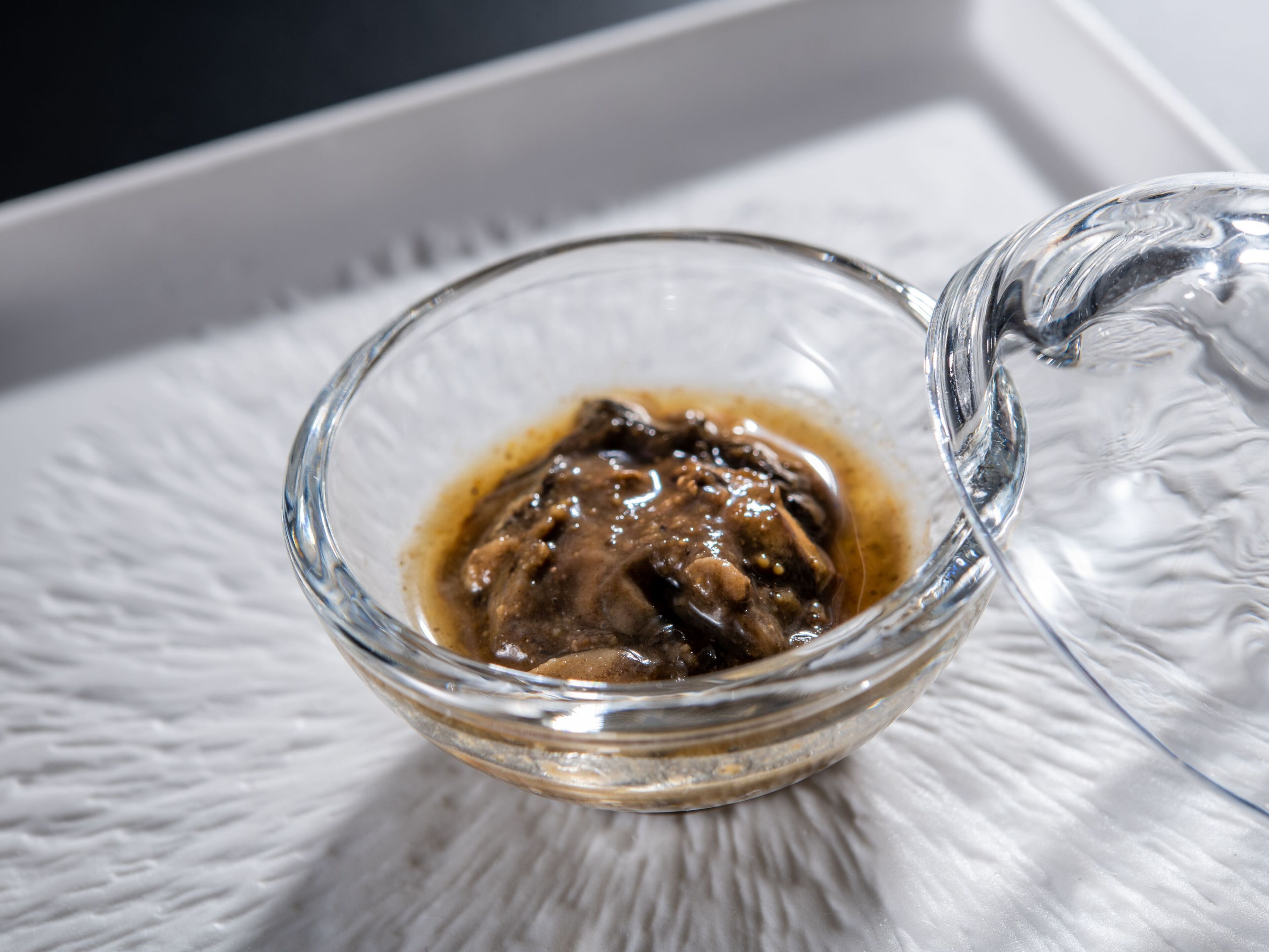 Un bol en verre transparent contenant une substance brun foncé, posé sur une surface blanche texturée.