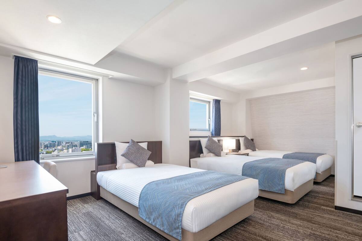 A hotel room with two beds, a desk, and a large window offering a city view.