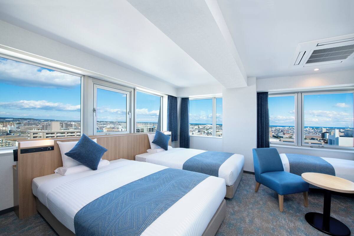 A hotel room with two beds, a chair, a small table, and large windows offering a city view.