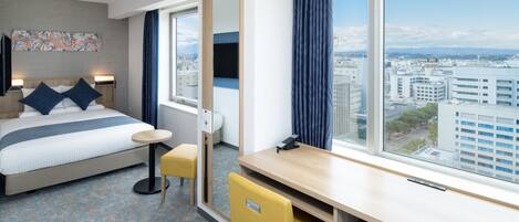 A hotel room with a bed, a desk, a chair, a large window with a city view, and a small table.
