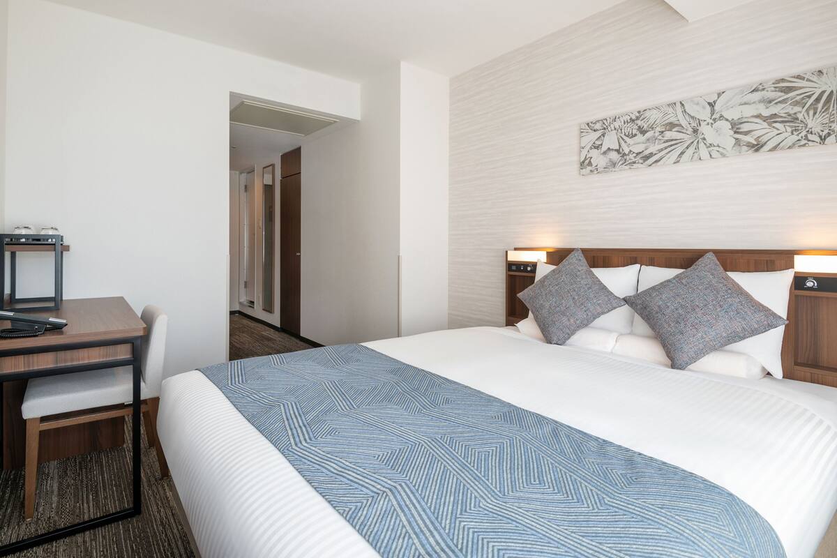A hotel room with a bed, a desk, and a chair. There is a door leading to another room.