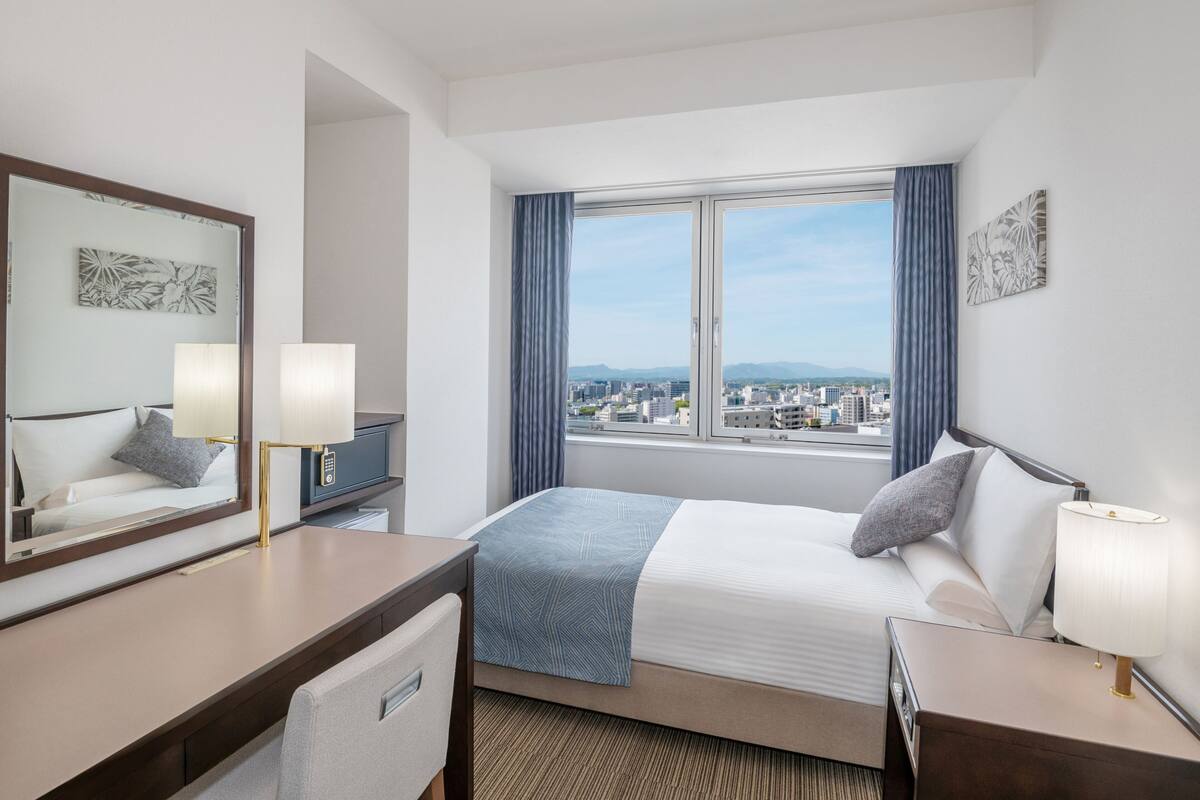 A hotel room with a bed, a desk, a chair, a lamp, a mirror, and a window with a city view.