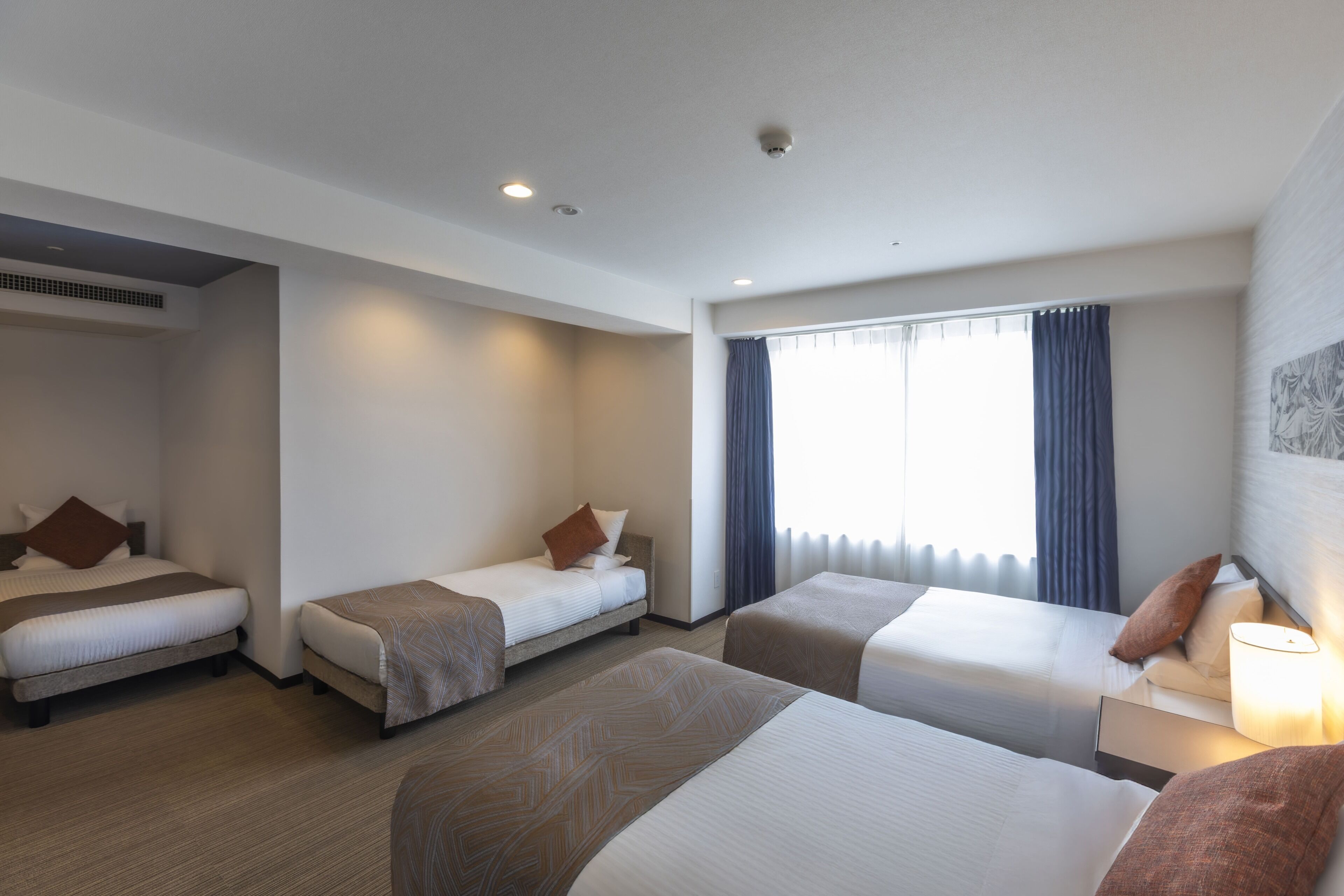 A hotel room with three beds, a large window with curtains, and a ceiling-mounted security camera.