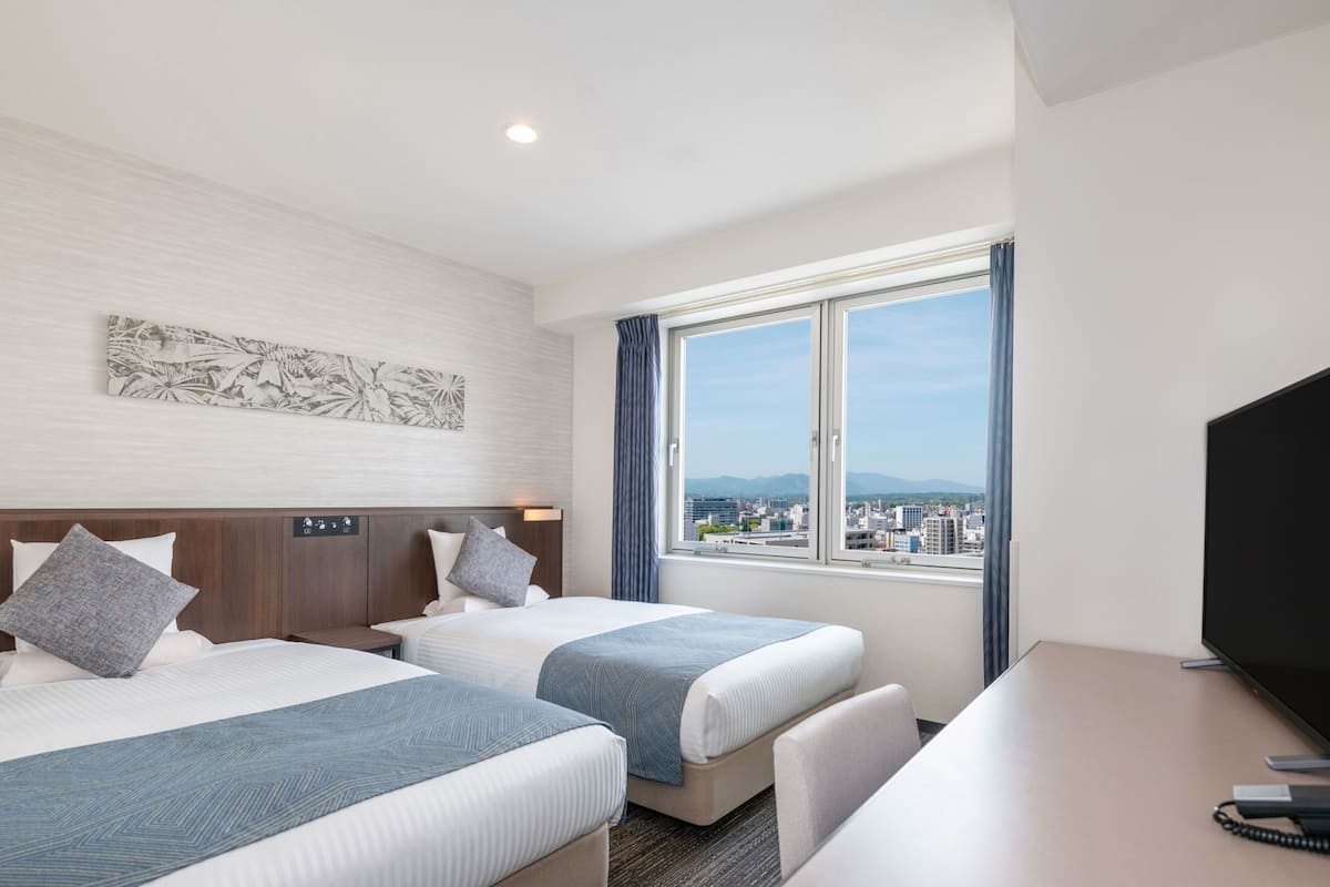 A hotel room with two beds, a desk, a television, and a window with a city view.