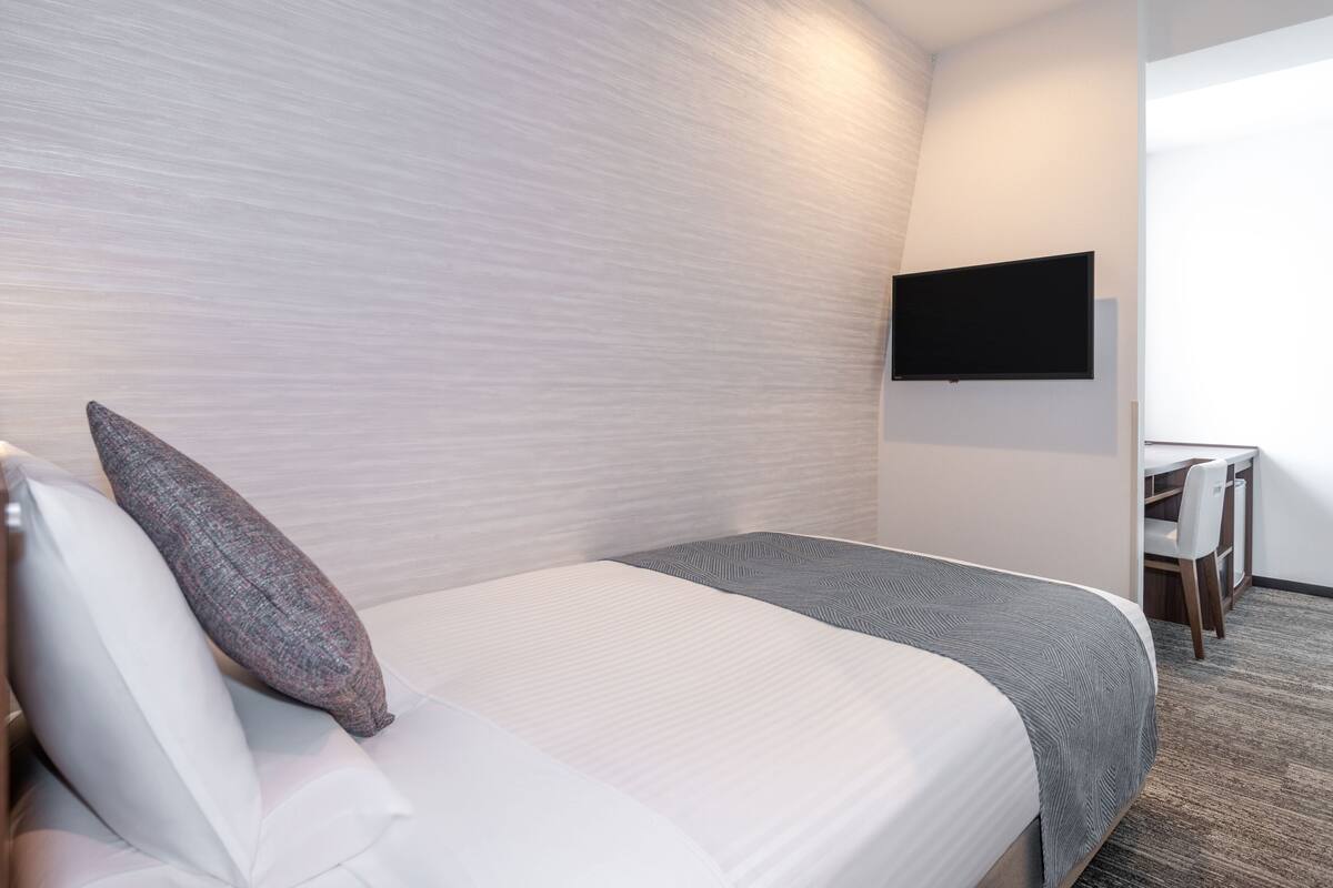 A hotel room with a bed, pillows, a TV mounted on the wall, and a desk with a chair.