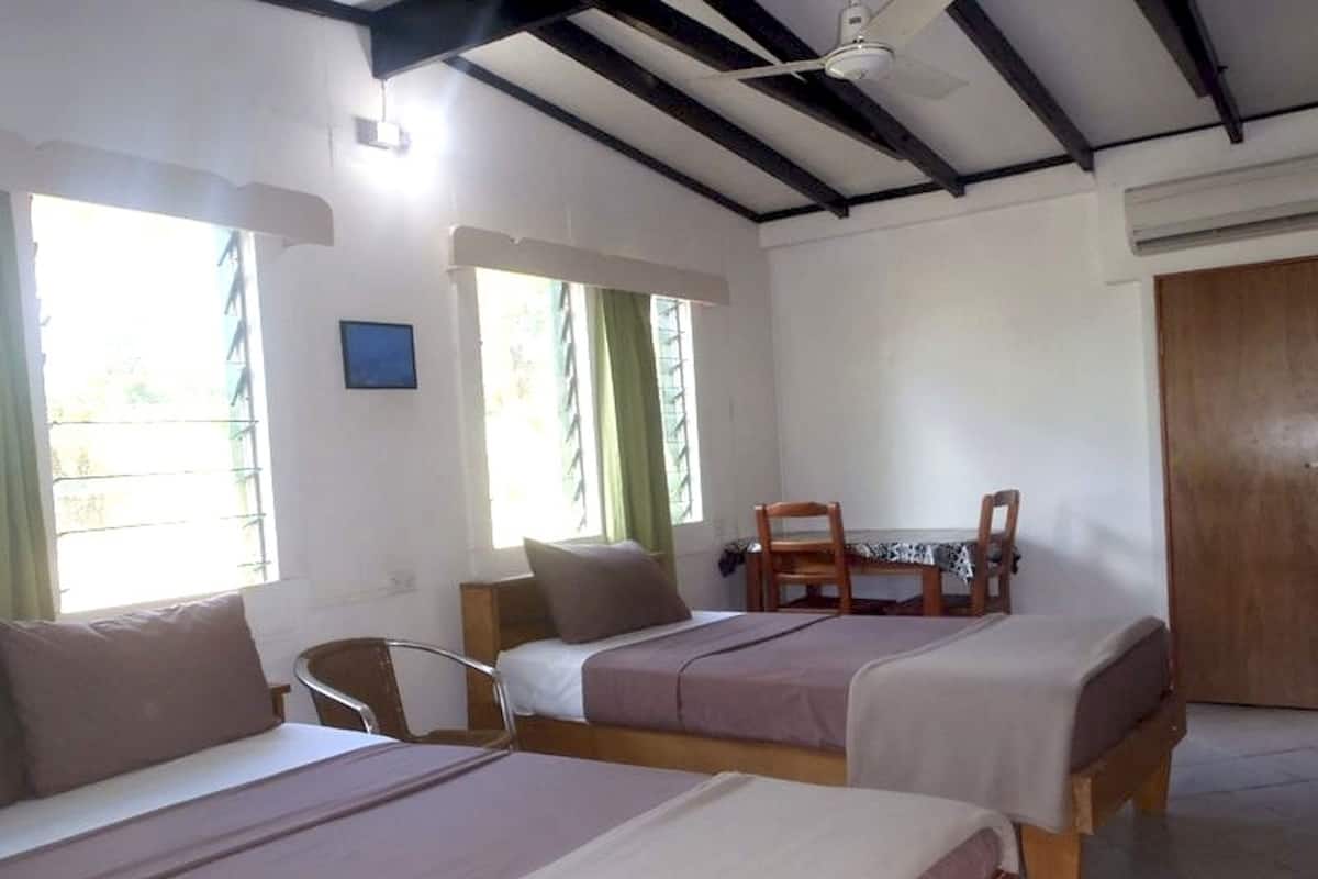 A room with two single beds, a wooden table with chairs, a window with curtains, and a ceiling fan.