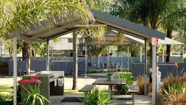 A covered outdoor seating area with a table and chairs, surrounded by plants and a fenced area.