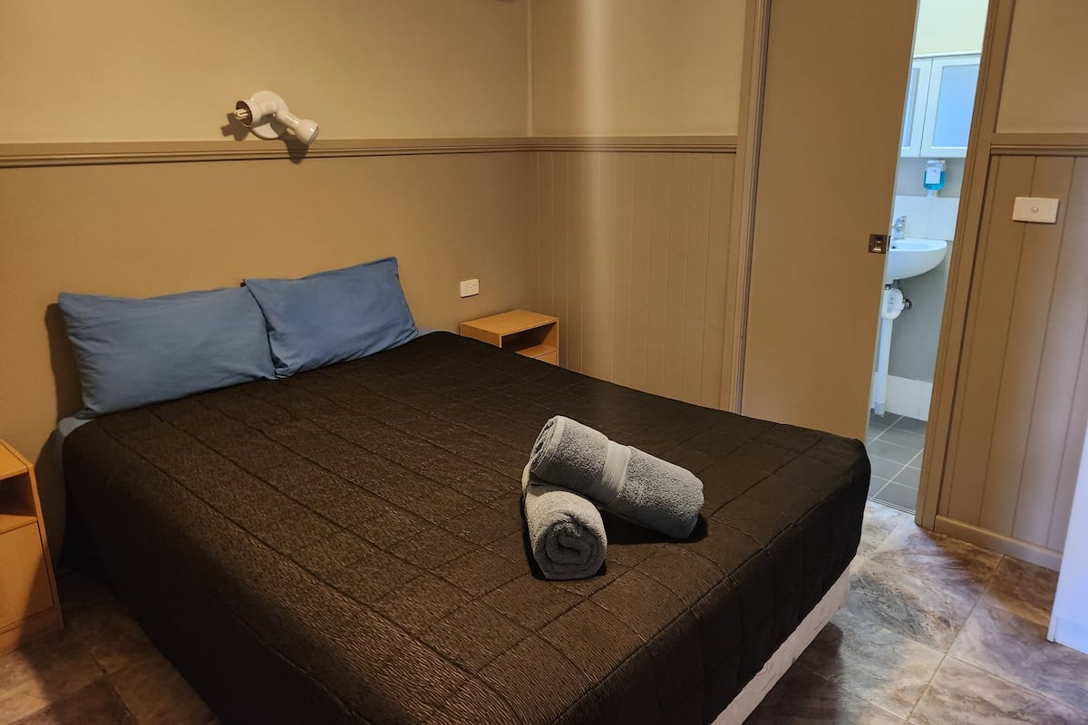 Ein ordentlich bezogenes Bett mit einer dunkelbraunen Decke, zwei blauen Kissen und einem aufgerollten Handtuch.